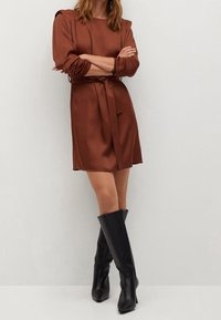 Brązowa satynowa sukienka z paskiem, z długimi rękawami i kwadratowym dekoltem, połączona z czarnymi dopasowanymi botkami do kolan.