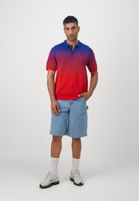Punane ja sinine gradientiga polo särk, millel on krae, koos heleda teksashortsiga ja valgete tennisekingadega hallide detailidega.