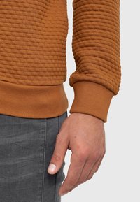 Gros plan de la main d’une personne à côté de sa hanche, portant un haut marron texturé à manches longues et un pantalon gris foncé sur un fond uni.