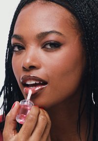 Przezroczysty błyszczyk do ust w małym słoiczku, aplikowany za pomocą gąbeczki, o błyszczącym wykończeniu i lekko różowym odcieniu. Długie, splecione warkocze.