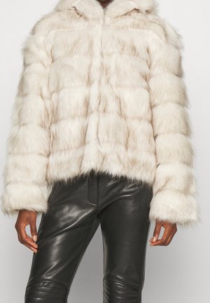 Persoon draagt een crèmekleurige nep bontjas met lange mouwen en zwarte leren broek, staand met armen ontspannen langs het lichaam.