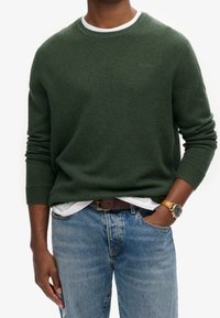 Pull en tricot vert foncé avec un col rond blanc, porté par-dessus une chemise blanche, associé à un jean bleu clair et une ceinture brune.