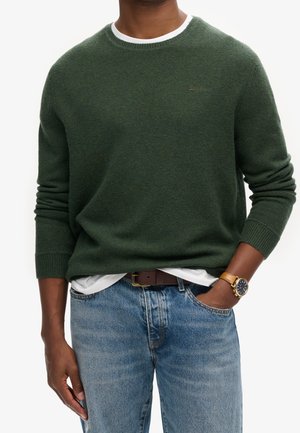 Pull en tricot vert foncé avec un col rond blanc, porté par-dessus une chemise blanche, associé à un jean bleu clair et une ceinture brune.