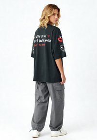 Melna pārspīlēta T-krekls ar grafiskiem uzrakstiem un dizainiem, īsām piedurknēm, kopā ar brīviem pelēkiem kravas biksēm un baltām sporta apaviem.