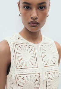 Crochet top in off-white with circular motifs and open stitch patterns. Features a round neckline and a button closure at the front.