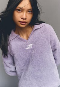 Lila, flauschiger Pullover mit Kragen, ausgestattet mit einem weißen Logo auf der Brust und langen, leicht übergroßen Ärmeln. Weicher, strukturierter Stoff.