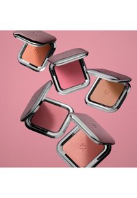 Vijf vierkante blushcompacts met glanzende zilveren randen, met verschillende tinten perzik en roze in een zachte, matte textuur tegen een roze achtergrond.
