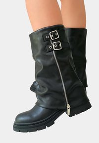 Bottes noires en cuir montantes avec un design décontracté, comportant des éléments en métal argenté, deux accents de boucle et une fermeture éclair sur une semelle texturée.