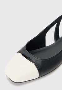 Zapato plano en blanco y negro con parte superior texturizada negra y puntera blanca lisa. Presenta un diseño con recortes para mayor transpirabilidad.