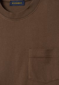 T-shirt di cotone marrone con scollatura tonda, maniche corte e taschino sul lato sinistro del petto. Presenta un'etichetta cucita con la scritta "PIOMBO" in blu e giallo.