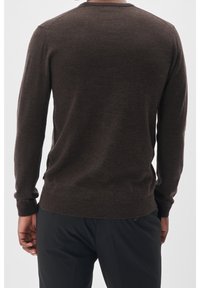 Man som bär en åtsittande mörkbrun stickad tröja med lång ärm och svarta byxor, sedd bakifrån mot en enkel bakgrund.