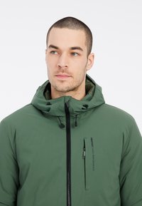 Chaqueta verde impermeable con cremallera frontal, capucha ajustable y bolsillo en el pecho. Textura suave con un acabado mate y detalle del logotipo en el lado.