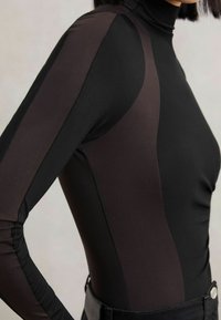 Top negru cu mânecă lungă, realizat dintr-un material neted, cu panouri mai închise la culoare și un design cu guler înalt, asociat cu blugi negri.