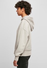 Hoodie grigio chiaro con cappuccio regolabile, polsini e fondo a costine. Presenta una vestibilità rilassata e una texture morbida, indossato con pantaloni neri.