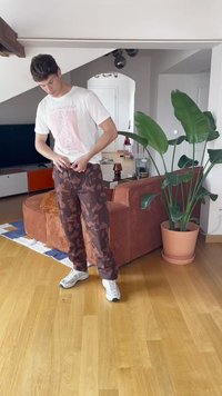 Burgundy toņu kamuflāžas bikses ar kabatām, apvienotas ar baltu grafisko t-kreklu un baltām sporta kurpēm, stāvot uz koka grīdas.