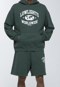 Conjunto de sudadera con capucha y pantalones cortos en verde oscuro que presenta un gráfico blanco "LOWLIGHTS WORLDWIDE" con un mapa mundial. Material suave, mezcla de algodón.