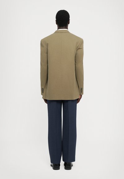 Brauner maßgeschneiderter Blazer mit strukturierten Schultern, kombiniert mit navyblauen Hosen. Glatte Stofftextur, schlichtes Design und Ein-Knopf-Verschluss.