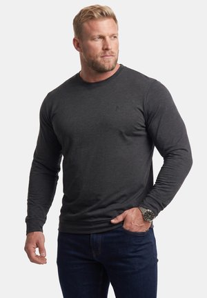 Uomo biondo con la barba che indossa una camicia scura a maniche lunghe, jeans scuri e un orologio da polso in metallo, guarda a sinistra con una mano in tasca.