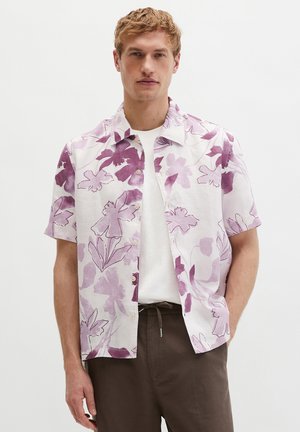 Mann trägt weißes Kurzarmhemd mit lila Blumenmuster, weißes T-Shirt und braune Hose, steht vor neutralem Hintergrund.