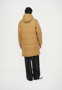 Siltuma brūnas pūkainais mētelis ar kapuci, quilted dizains un vidēja garuma griezums. Kombinēts ar brīva piegriezuma melna raibā auduma biksēm.