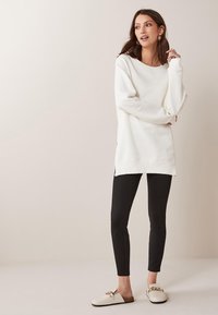 Sudadera blanca de manga larga con un corte relajado, aberturas laterales, combinada con leggins negros y zapatos tipo slip-on color crema con un detalle de cadena dorada.