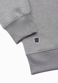 Grijze sweater met geribbelde mouwen en onderkant. Heeft een klein vierkant logo-patch nabij de mouw. Zacht, textuurig stof.