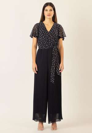 Schwarzer Jumpsuit mit einem durchsichtigen Overlay aus Pünktchenmuster, plissiertem weitem Bein design und einem Stoffbinder in der Taille. Kurze, schwingende Ärmel, glatte Textur.