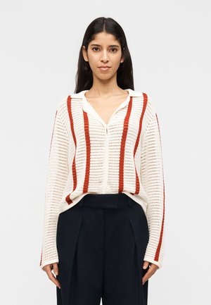 Žena s dlhými tmavými vlasmi nosí biely pletený sveter s vertikálnymi hrdzavými pruhmi a voľné čierne nohavice s vysokým pásom, stojaca na bielom pozadí.