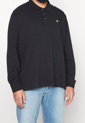 Mężczyzna ubrany w czarną koszulkę polo z długim rękawem z żółtym logiem ptaka na piersi oraz jasnoniebieskie dżinsy, stojący na jednolitym tle.