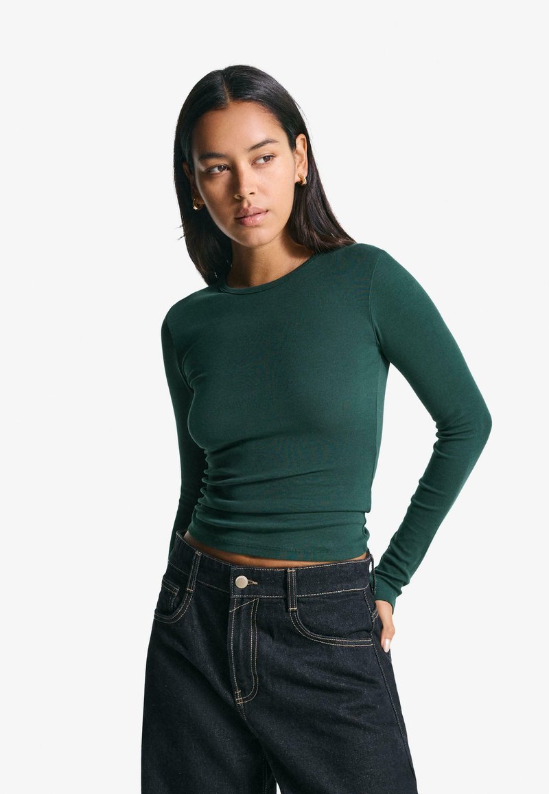 Langärmliges, dunkelgrünes Ripp-Top mit figurnahem Schnitt und Rundhalsausschnitt, kombiniert mit hoch taillierten, dunklen Jeans.