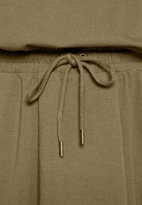 Oliwkowa bluzka o luźnym kroju, z centralnym sznurkiem ze metalowymi końcówkami i gładkim, teksturowanym materiałem.