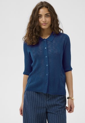 Göndör barna hajú nő, kék rövid ujjú, gombos kötött felsőt és kék, vékony csíkos nadrágot visel, egyszínű háttér előtt áll.