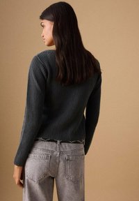 Dunkelgrüner, gerippter Pullover mit langen Ärmeln, kombiniert mit hellgrauen Jeans mit Gesäßtaschen und einem schwarz mit Nieten besetzten Gürtel.