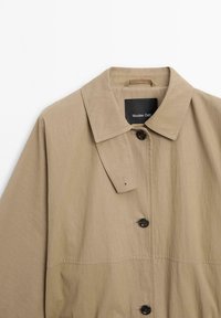 Manteau léger beige avec un col, fermeture à bouton et une texture lisse. Détails notables : une étiquette et un bouton unique.