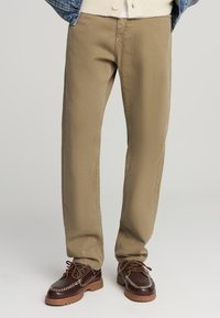 Pantaloni khaki con un design a gamba dritta, realizzati in resistente tessuto di cotone, caratterizzati da cuciture minime e tasche discrete, abbinati a scarpe marroni.