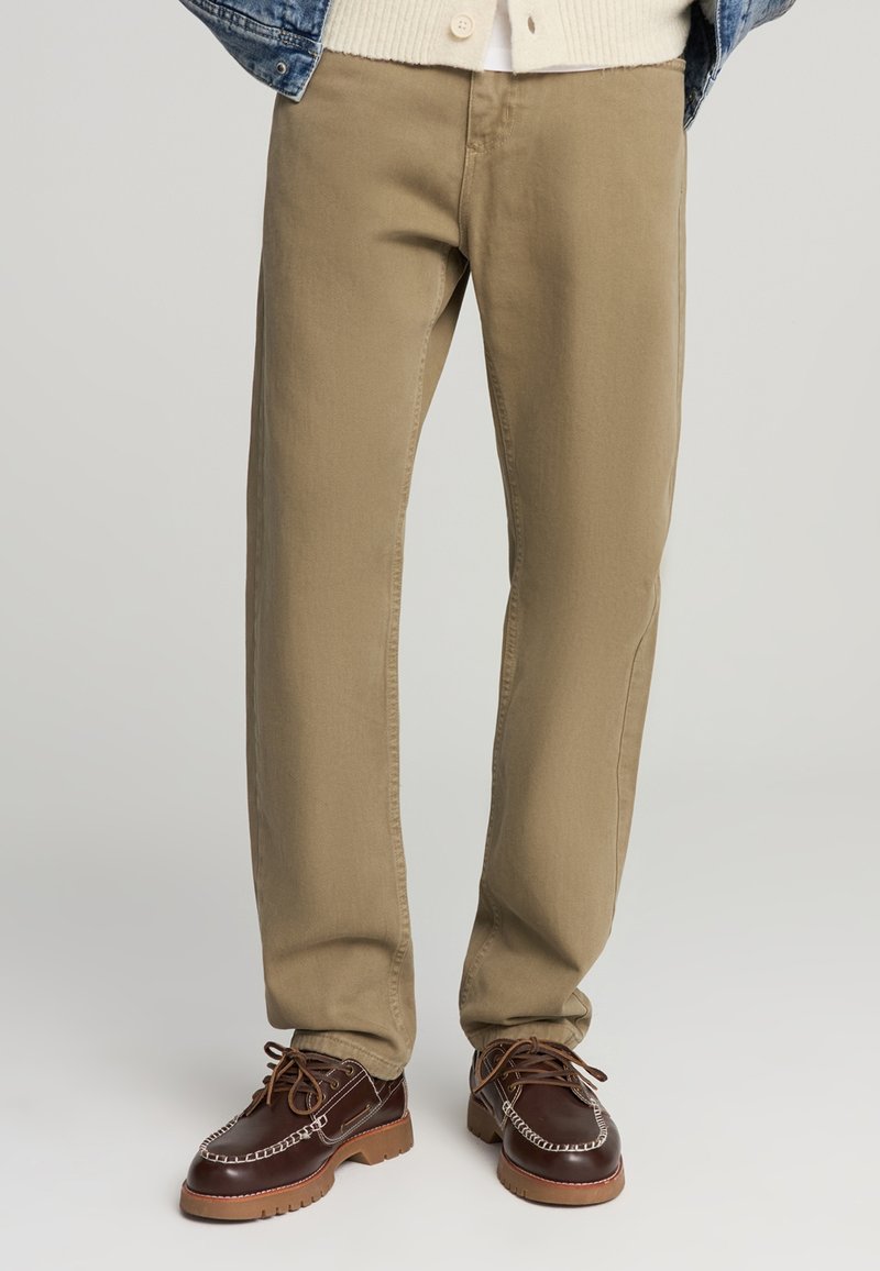Pantaloni khaki con un design a gamba dritta, realizzati in resistente tessuto di cotone, caratterizzati da cuciture minime e tasche discrete, abbinati a scarpe marroni.