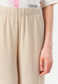 Beiga texturerade byxor med elastiskt midjeband, med en rynkad tygdesign. Hand delvis synlig på sidan.