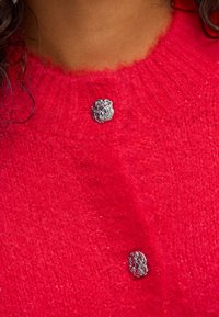 Gros plan sur un pull en laine rouge avec deux boutons en argent texturés près du col, porté sur la partie supérieure de la poitrine d'une personne.