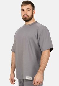 T-shirt grigio chiaro a maniche corte con collo a giro, caratterizzato da dettagli di cucitura e un'etichetta di marca in basso a sinistra. Texture del tessuto morbida.