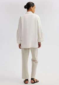 Camicia aderente color crema con spalle scese e polsini con bottoni, abbinata a pantaloni beige chiaro su misura e sandali neri con cinturini.