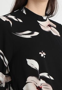 Blouse noire avec un motif floral en crème et rouge. Présente un col haut et un tissu léger, avec des manches fluides et une texture douce.