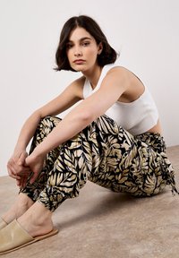 Pantalons harem à motif noir et doré avec des poignets élastiques et un top court blanc. Modèle assis sur une surface de sol marron clair.