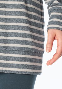 Randig långärmad topp i beige och marinblå, med mjuk textur och avslappnad passform, matchad med turkosa leggings. Handen vilar bredvid fållen.
