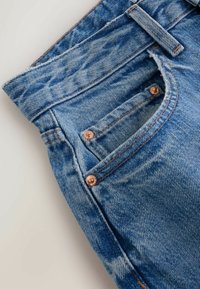 Gros plan sur la poche d'un jean en denim bleu avec des rivets en cuivre, une passants de ceinture visible et des coutures détaillées sur un tissu délavé.