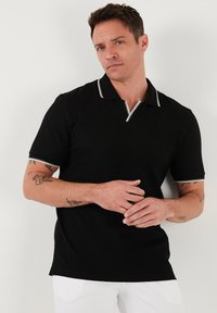 Černé polo tričko vyrobené z texturované látky, s zipovým předním dílem a pruhovaným límcem. Krátké rukávy s odpovídajícími pruhovanými detaily.