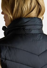 Blonde sieviete redzama no aizmugures, tērpta melnā dūnu jakā ar augstu apkakli un izšūtu "Joules" logo uz muguras.