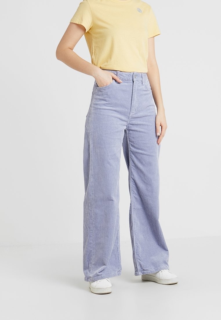 Personne portant un pantalon large en velours côtelé violet clair, une chemise jaune à manches courtes et des baskets blanches, une main dans la poche.