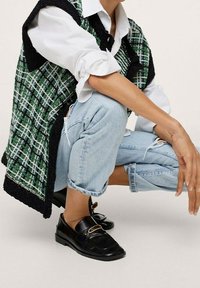 Grünes und schwarzes gewebtes Weste, die über einem weißen Shirt getragen wird, kombiniert mit hellblauen, zerrissenen Jeans und schwarzen Slippern mit goldenen Hardware-Details.