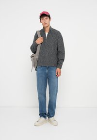Suéter gris de punto texturizado con media cremallera, jeans de mezclilla azules y zapatillas blancas. Los accesorios incluyen una gorra rosa y un bolso gris de hombro.