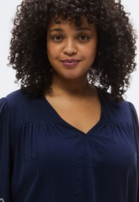 Haut bleu marine à col en V en tissu léger avec des détails plissés aux épaules, présentant une texture douce et une coupe décontractée.